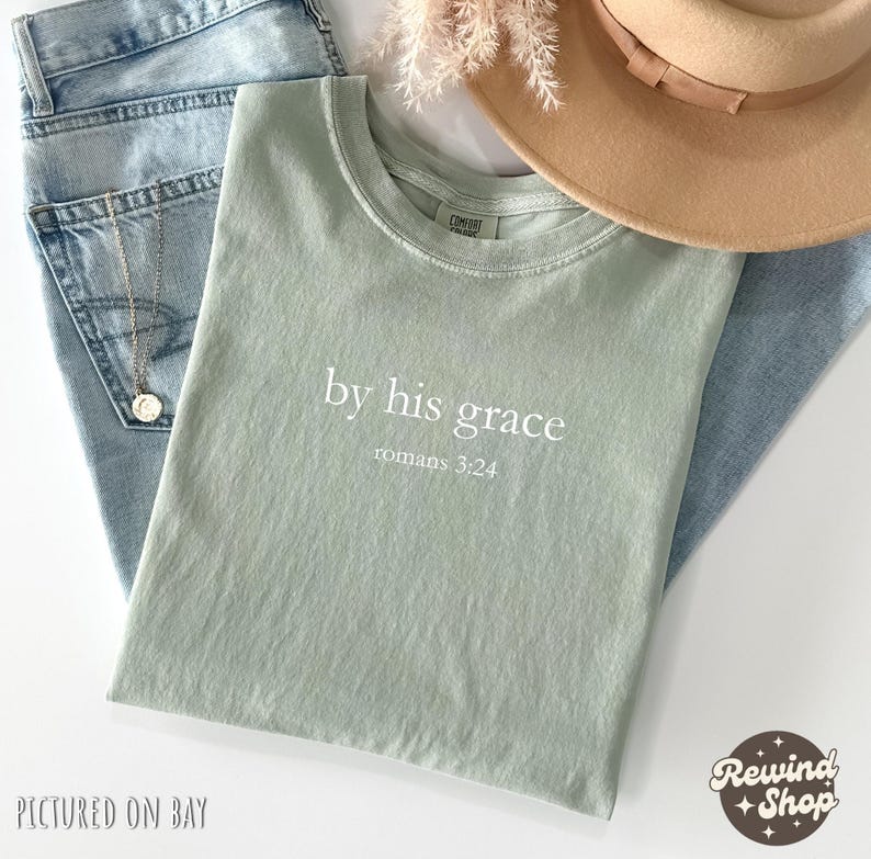 May include: A sage green t-shirt with the text "by his grace romans 3:24" in white. The shirt is laid on top of light blue jeans, with a gold necklace and a tan hat. The image is from Rewind Shop.