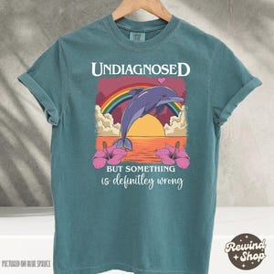 Op de afbeelding: Tealblauw T-shirt met een afbeelding van een dolfijn die uit het water springt met een regenboog en roze bloemen. De tekst "Undiagnosed but something is definitely wrong" staat op het shirt gedrukt.