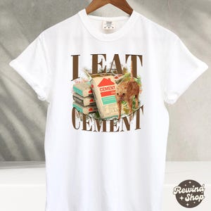 May include: White t-shirt with a brown graphic that says "I EAT CEMENT" and features a cartoon cat eating bags of cement.