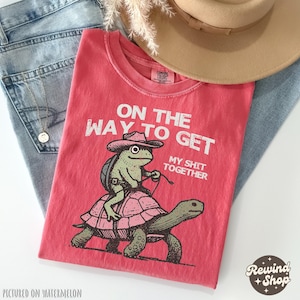May include: Red t-shirt with a graphic of a frog riding a turtle. The text on the shirt reads "On the way to get my shit together".