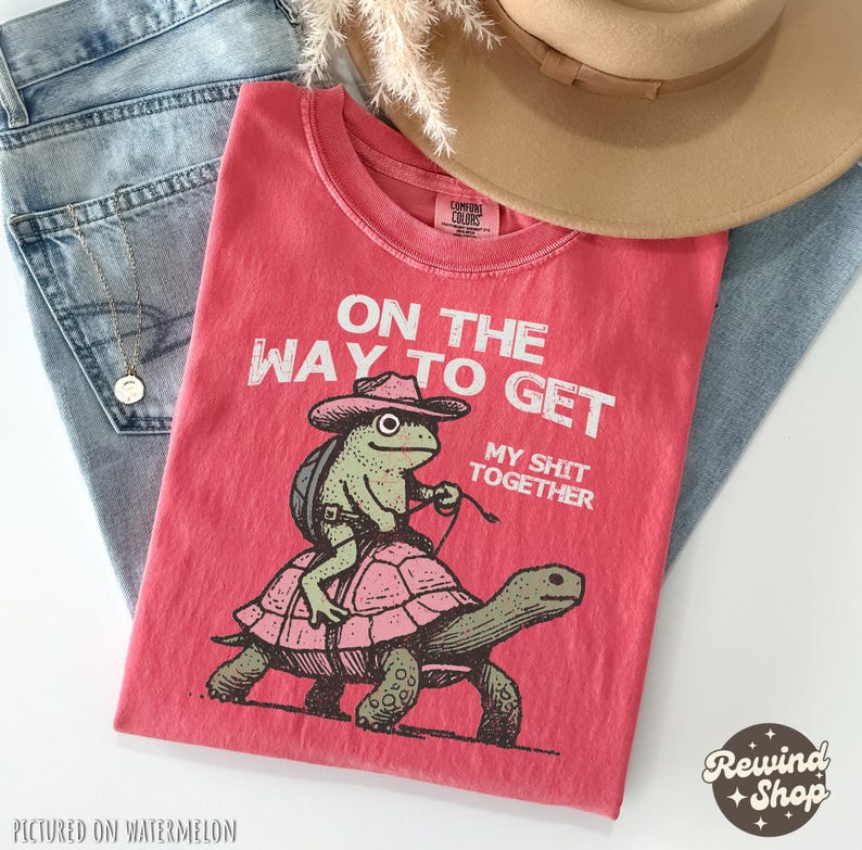 May include: Red t-shirt with a graphic of a frog riding a turtle. The text on the shirt reads "On the way to get my shit together".