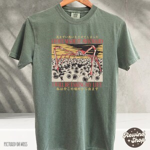 May include: A moss green t-shirt featuring a graphic design with a red and beige crab-like creature with multiple eyes. The text "I HAVE MADE IT AWKWARD" and "I'LL BE TAKING MY LIFE" are also visible.