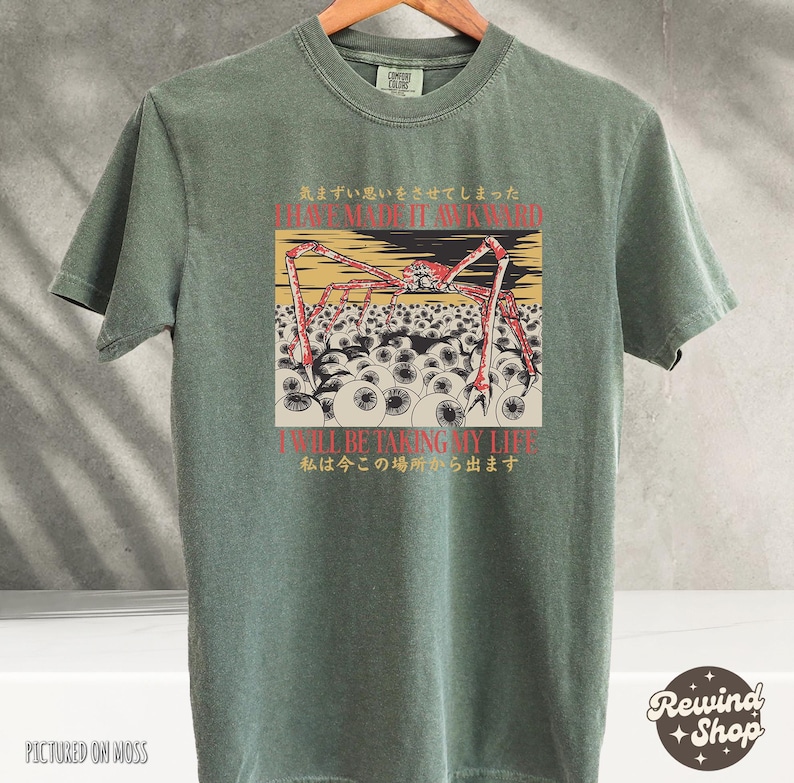 May include: A moss green t-shirt featuring a graphic design with a red and beige crab-like creature with multiple eyes. The text "I HAVE MADE IT AWKWARD" and "I'LL BE TAKING MY LIFE" are also visible.