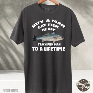 May include: A dark gray t-shirt with white text that reads "Buy a man eat fish, he day teach fish man to a lifetime".