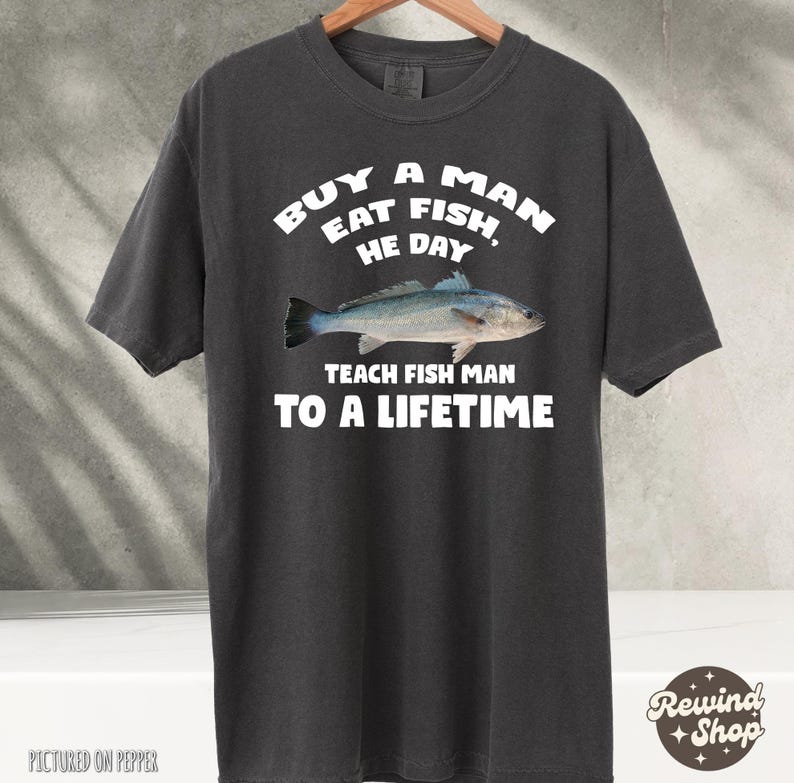 May include: A dark gray t-shirt with white text that reads "Buy a man eat fish, he day teach fish man to a lifetime".