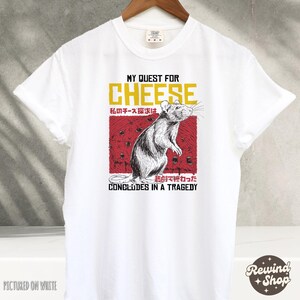 Op de afbeelding: Wit T-shirt met een afbeelding van een rat en de tekst "My Quest for Cheese" in rood en geel. De tekst "私のチーズ探求は悲劇で終わった" is in het Japans en betekent "Mijn zoektocht naar kaas eindigde in een tragedie".
