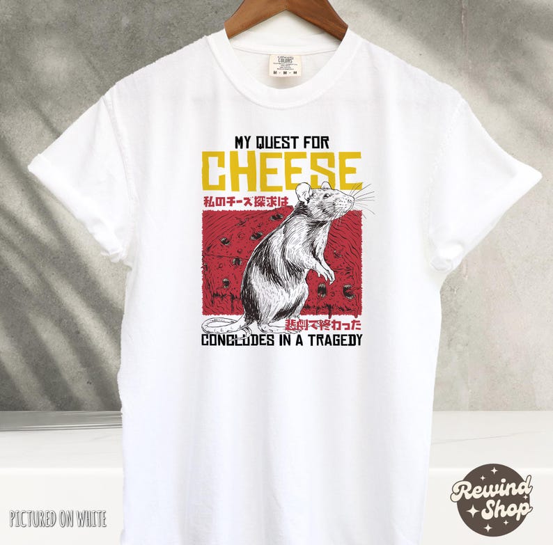 Op de afbeelding: Wit T-shirt met een afbeelding van een rat en de tekst "My Quest for Cheese" in rood en geel. De tekst "私のチーズ探求は悲劇で終わった" is in het Japans en betekent "Mijn zoektocht naar kaas eindigde in een tragedie".