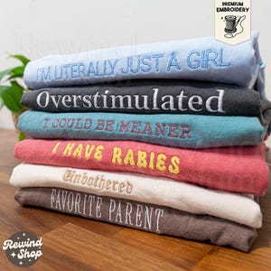 May include: A stack of six folded t-shirts in light blue, dark grey, teal, coral, white, and brown. Each shirt features embroidered text: I'M LITERALLY JUST A GIRL, Overstimulated, I COULD BE MEANER, I HAVE RABIES, Unbothered, and FAVORITE PARENT.