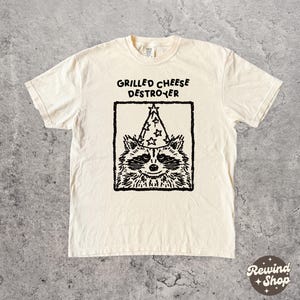 May include: Cream-colored t-shirt with a black graphic of a raccoon wearing a wizard hat, inside a black square. The text "GRILLED CHEESE DESTROYER" is above the graphic. The shirt is on a gray surface.