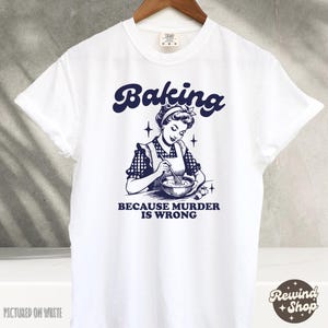 May include: White t-shirt with a blue graphic of a woman baking. The text on the shirt reads "Baking because murder is wrong."