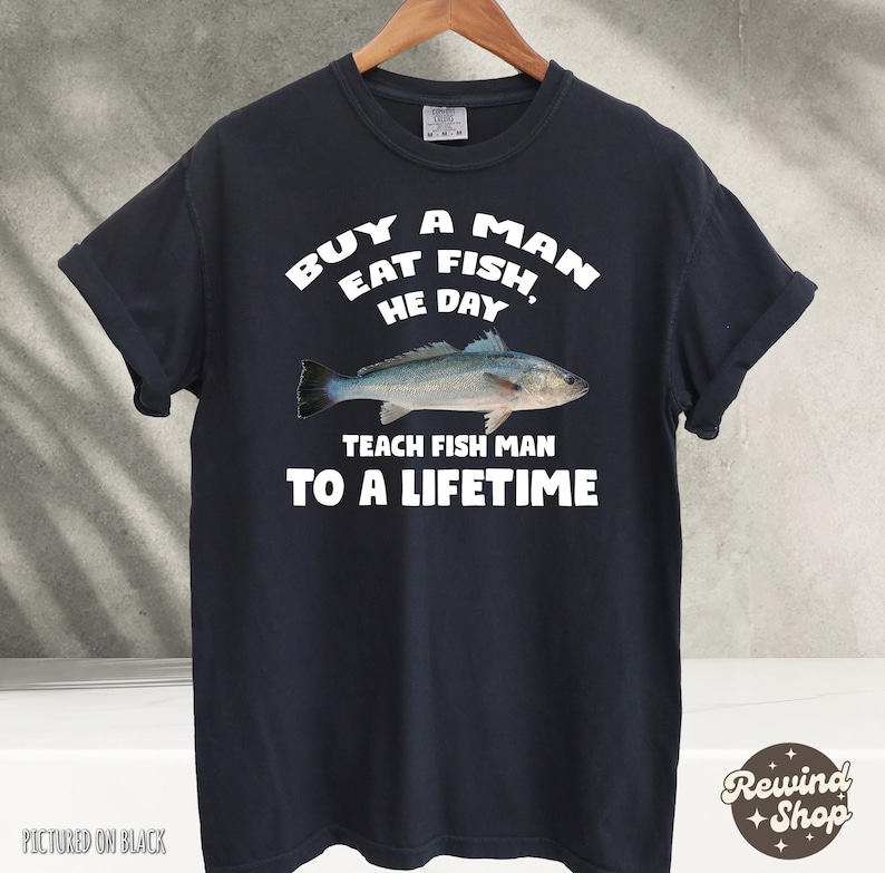 May include: A black t-shirt with white text that reads "Buy a man eat fish, he day teach fish man to a lifetime". The shirt features a graphic of a fish.