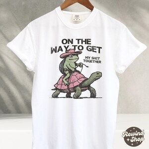 May include: White t-shirt with a graphic of a frog riding a turtle. The text on the shirt reads "On the way to get my shit together."