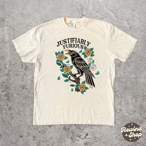 Puede incluir: Camiseta color crema con un diseño de un cuervo negro posado en una rama, rodeado de rosas amarillas y hojas verdes. El texto "JUSTIFIABLY FURIOUS" está arqueado sobre el cuervo. El diseño es de inspiración vintage.