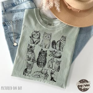 K&ouml;nnte beinhalten: Hellgr&uuml;nes T-Shirt mit einer schwarzen Illustration verschiedener Katzenrassen. Das Shirt ist auf einer blauen Jeans gefaltet, mit einer Halskette und einem beigen Hut. Der Text "PICTURED ON BAY" befindet sich unten links und "Rewind Shop" unten rechts.