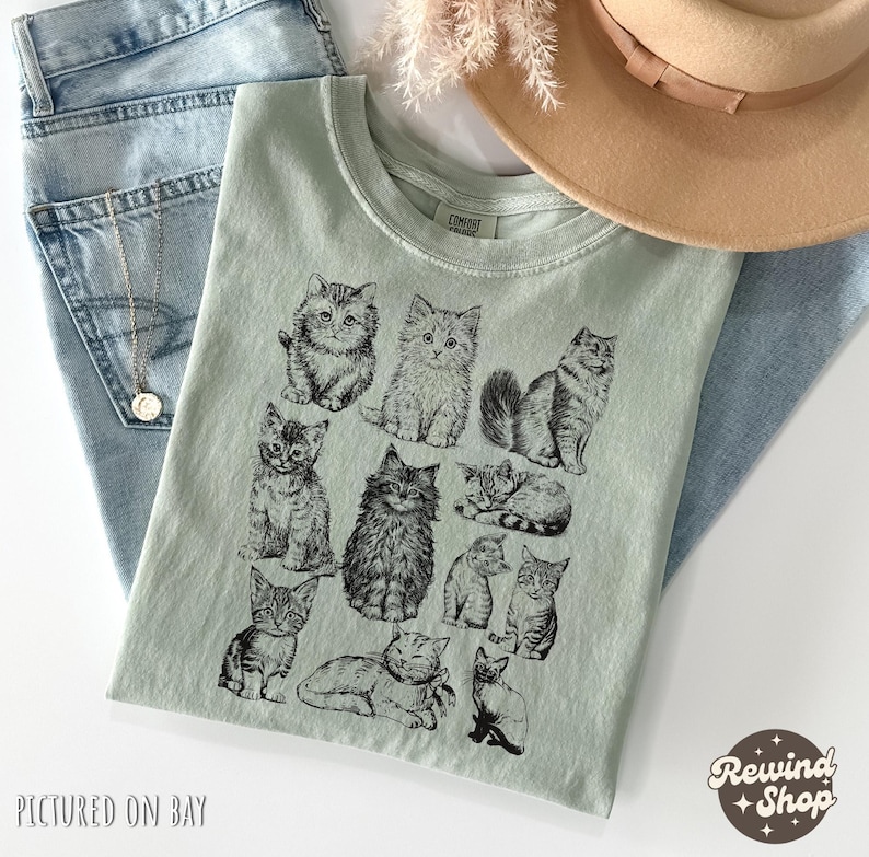 K&ouml;nnte beinhalten: Hellgr&uuml;nes T-Shirt mit einer schwarzen Illustration verschiedener Katzenrassen. Das Shirt ist auf einer blauen Jeans gefaltet, mit einer Halskette und einem beigen Hut. Der Text "PICTURED ON BAY" befindet sich unten links und "Rewind Shop" unten rechts.