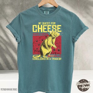 Op de afbeelding: Tealblauwe T-shirt met een afbeelding van een rat en de tekst "My Quest for Cheese Concludes in a Tragedy".