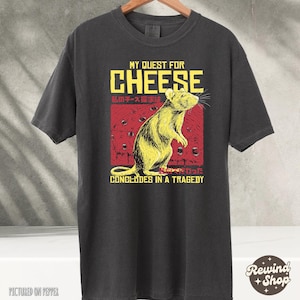 Op de afbeelding: Een zwart t-shirt met een afbeelding van een rat en de tekst "My Quest for Cheese concludes in a tragedy". De tekst is in het Engels en Japans.