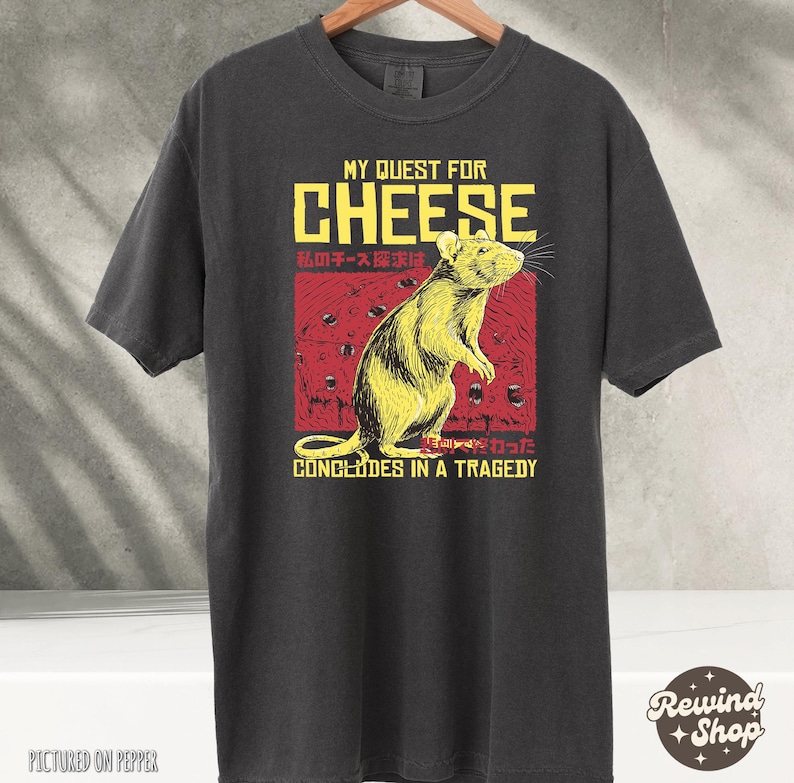 Op de afbeelding: Een zwart t-shirt met een afbeelding van een rat en de tekst "My Quest for Cheese concludes in a tragedy". De tekst is in het Engels en Japans.