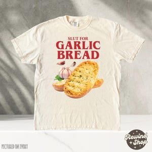 Puede incluir: Una camiseta blanca con el texto "Slut for Garlic Bread" en letras rojas. La camiseta presenta un gráfico de dos rebanadas de pan de ajo con un diente de ajo.