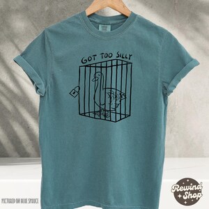 May include: A teal blue t-shirt with a black graphic of a goose in a cage. The text "Got Too Silly" is above the graphic.