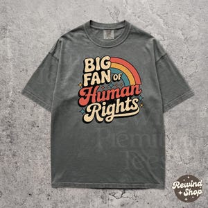 Puede incluir: Camiseta gris con el texto "BIG FAN of Human Rights" en un diseño retro de arcoíris. La camiseta tiene mangas cortas y cuello redondo. La tela es suave y cómoda.