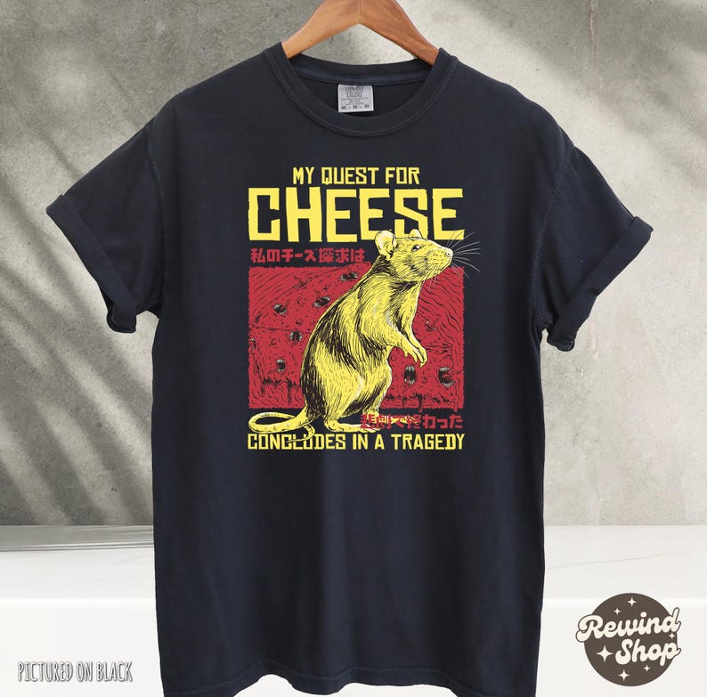 Op de afbeelding: Zwart T-shirt met een afbeelding van een rat en de tekst "My Quest for Cheese" in rood en geel. De tekst "私のチーズ探求は" is in het Japans en de tekst "CONCLUDES IN A TRAGEDY" is in zwart.