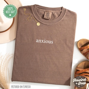 May include: A brown t-shirt with the word "anxious" printed in white on the front. The shirt is pictured on an espresso background.
