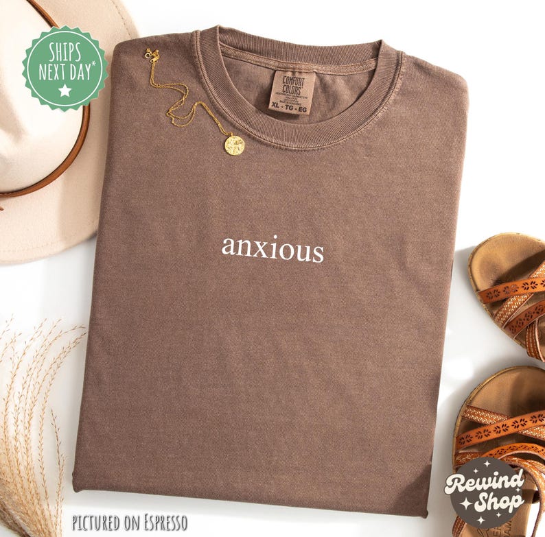 May include: A brown t-shirt with the word "anxious" printed in white on the front. The shirt is pictured on an espresso background.