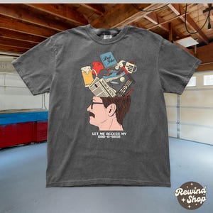 May include: Dark gray t-shirt featuring a graphic of a man's head with various items emerging from it, including a beer, a book titled "Dad Jokes", and a remote control. The text "LET ME ACCESS MY DAD-A-BASE" is below the graphic.