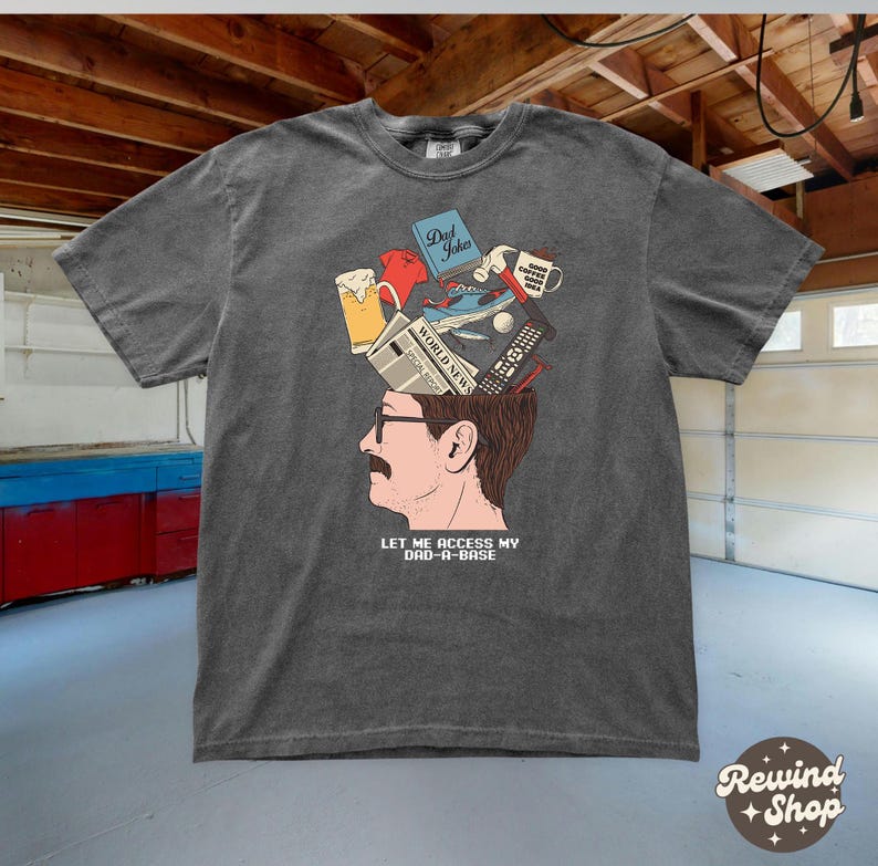 May include: Dark gray t-shirt featuring a graphic of a man's head with various items emerging from it, including a beer, a book titled "Dad Jokes", and a remote control. The text "LET ME ACCESS MY DAD-A-BASE" is below the graphic.