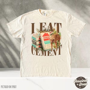 May include: A beige t-shirt with the text "I EAT CEMENT" printed on the front. The text is in brown letters with a brown outline. There is a picture of a cat eating a bag of cement. The cat is orange and white. The t-shirt is pictured on an ivory background.