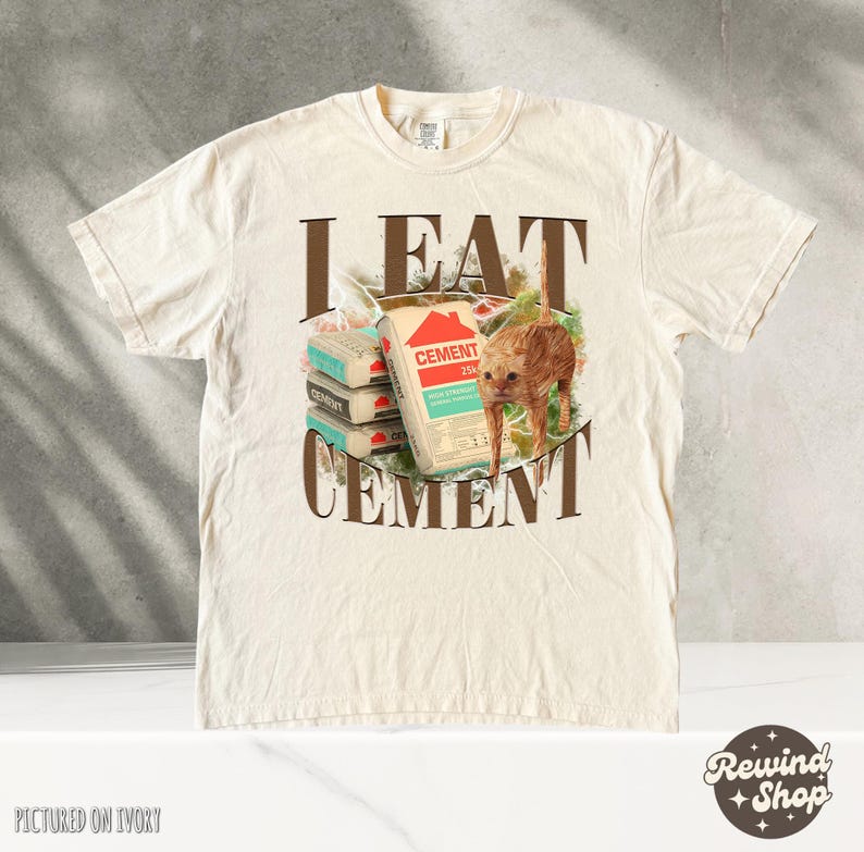 May include: A beige t-shirt with the text "I EAT CEMENT" printed on the front. The text is in brown letters with a brown outline. There is a picture of a cat eating a bag of cement. The cat is orange and white. The t-shirt is pictured on an ivory background.