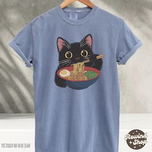 May include: A blue t-shirt with a black cat eating ramen noodles. The cat is wearing a red collar and has a bowl of ramen in front of it. The text "Rewind Shop" is in the bottom right corner of the image.