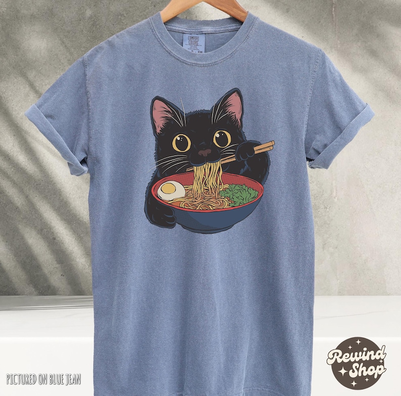 May include: A blue t-shirt with a black cat eating ramen noodles. The cat is wearing a red collar and has a bowl of ramen in front of it. The text "Rewind Shop" is in the bottom right corner of the image.