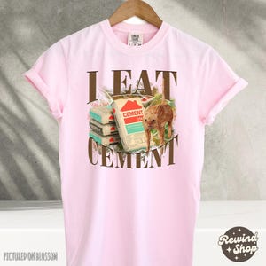 May include: Pink t-shirt with a brown graphic of a cat eating cement bags. The text "I EAT CEMENT" is printed on the shirt.