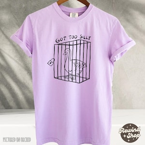 May include: A lavender t-shirt with the text "Got Too Silly" and a black and white illustration of a goose in a cage.