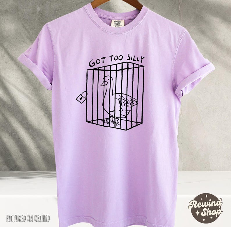May include: A lavender t-shirt with the text "Got Too Silly" and a black and white illustration of a goose in a cage.