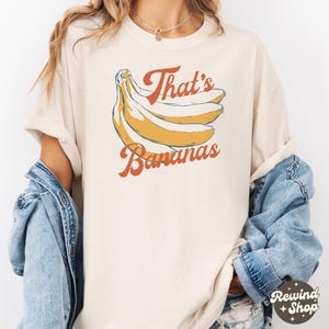 May include: Cream-colored t-shirt featuring a retro illustration of bananas and the text "That's Bananas" in orange script. The design includes a bunch of yellow bananas with blue outlines. The shirt has short sleeves and a relaxed fit.