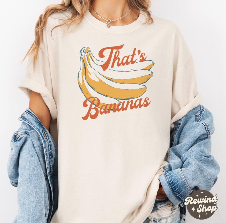 May include: Cream-colored t-shirt featuring a retro illustration of bananas and the text "That's Bananas" in orange script. The design includes a bunch of yellow bananas with blue outlines. The shirt has short sleeves and a relaxed fit.
