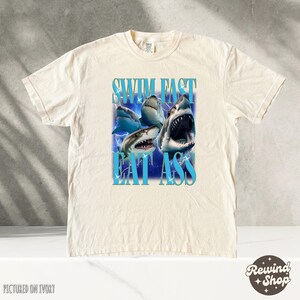 May include: A cream-colored t-shirt with a graphic of sharks and the text "Swim Fast Eat Ass".
