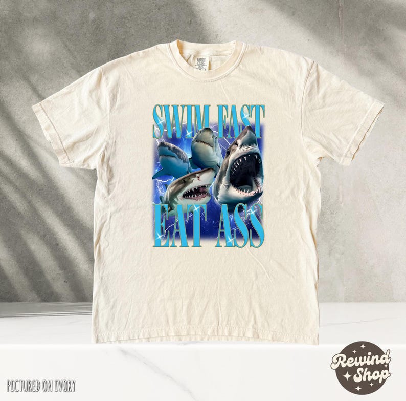 May include: A cream-colored t-shirt with a graphic of sharks and the text "Swim Fast Eat Ass".