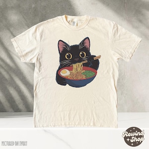 May include: A black cat with green eyes is eating ramen noodles from a bowl with chopsticks. The cat is wearing a white t-shirt with the text "Rewind Shop" printed on it. The t-shirt is pictured on an ivory background.