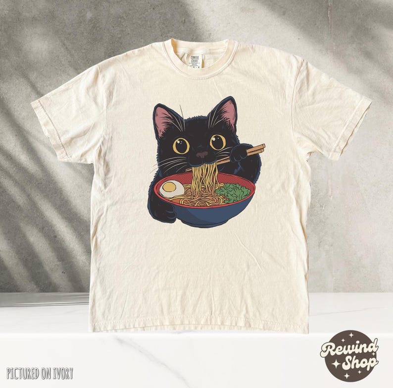 May include: A black cat with green eyes is eating ramen noodles from a bowl with chopsticks. The cat is wearing a white t-shirt with the text "Rewind Shop" printed on it. The t-shirt is pictured on an ivory background.