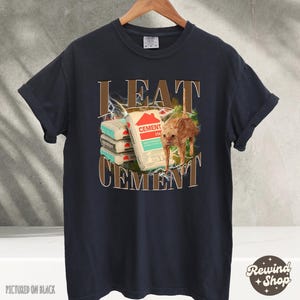 May include: A dark blue t-shirt with a graphic print of a cat eating cement bags. The text "I EAT CEMENT" is printed on the shirt.