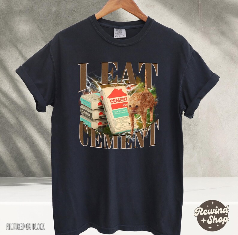 May include: A dark blue t-shirt with a graphic print of a cat eating cement bags. The text "I EAT CEMENT" is printed on the shirt.