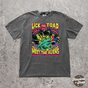 May include: Dark gray t-shirt with a colorful graphic. The graphic features a green frog with its tongue out, surrounded by stars and the text "Lick the Toad Meet the Aliens" in bright colors. The shirt has a vintage look.