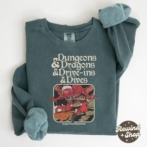 May include: Teal-colored sweatshirt with the text "Dungeons & Dragons & Drive-ins & Dives" in white. Below the text is a graphic of a red dragon wearing sunglasses, holding a plate of food, with the text "Escape From Flavortown".