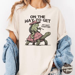 May include: A cream colored t-shirt with the text "On the way to get my shit together" and a cartoon frog riding a turtle.