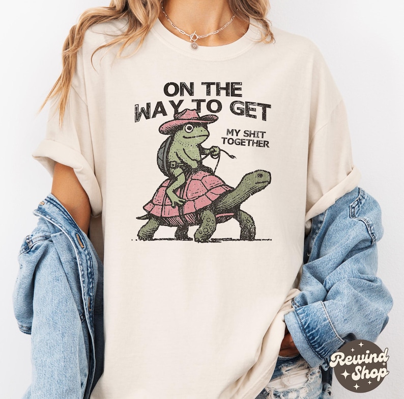 May include: A cream colored t-shirt with the text "On the way to get my shit together" and a cartoon frog riding a turtle.