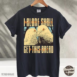 Op de afbeelding: Zwart T-shirt met een afbeelding van twee duiven met de tekst "I alone shall get this bread" in wit en geel.